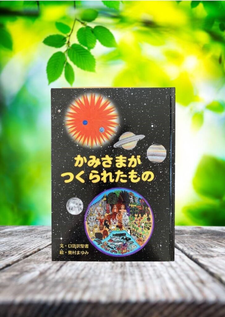 木のテーブルの上に絵本『かみさまがつくられたもの』が立てかけられ、宇宙といのちを描いた表紙が、緑にあふれる自然の背景に優しく映える