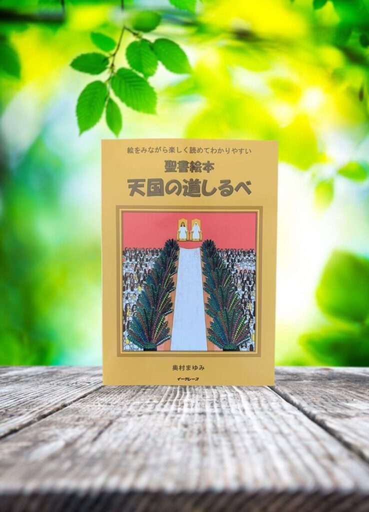 木のテーブルに立てかけられた絵本『天国の道しるべ』。背景には若葉が光に透ける緑の自然が広がり、やさしい雰囲気の中に本が静かに佇む